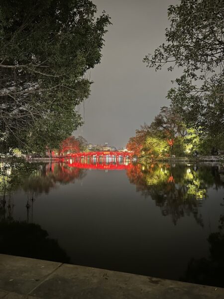 A nighttime view of Hoàn Kiếm Lake, reflecting the red lights of Huc Bridge, surrounded by lush trees.