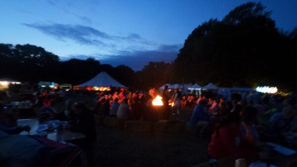 Crowds gathered around a big fire pit at night with glowing tents and food stalls in the background