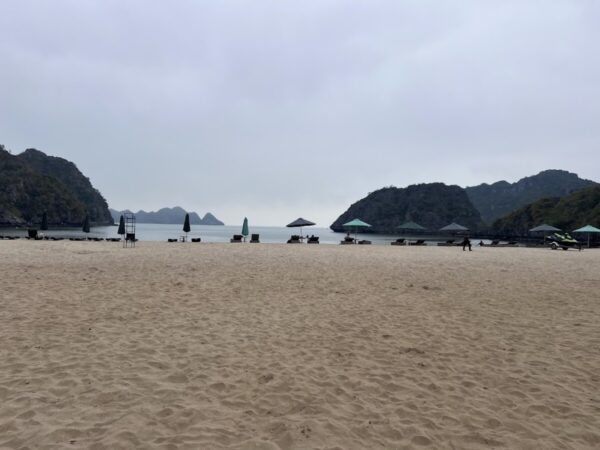 View of a sandy beach on Cat Ba Island, with umbrellas and deck chairs facing the calm sea and rocky islands in the background under a cloudy sky.